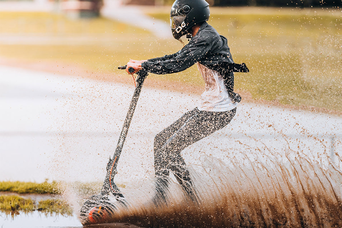 Man riding Inmotion Climber electric scooter through water and mud, wearing helmet and safety gear – showcasing off-road capability and waterproof design