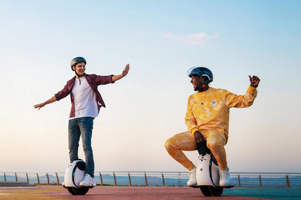 Two people wearing helmets riding the Inmotion E20 electric unicycles on a seaside path at sunset, smiling and balancing with their arms out.