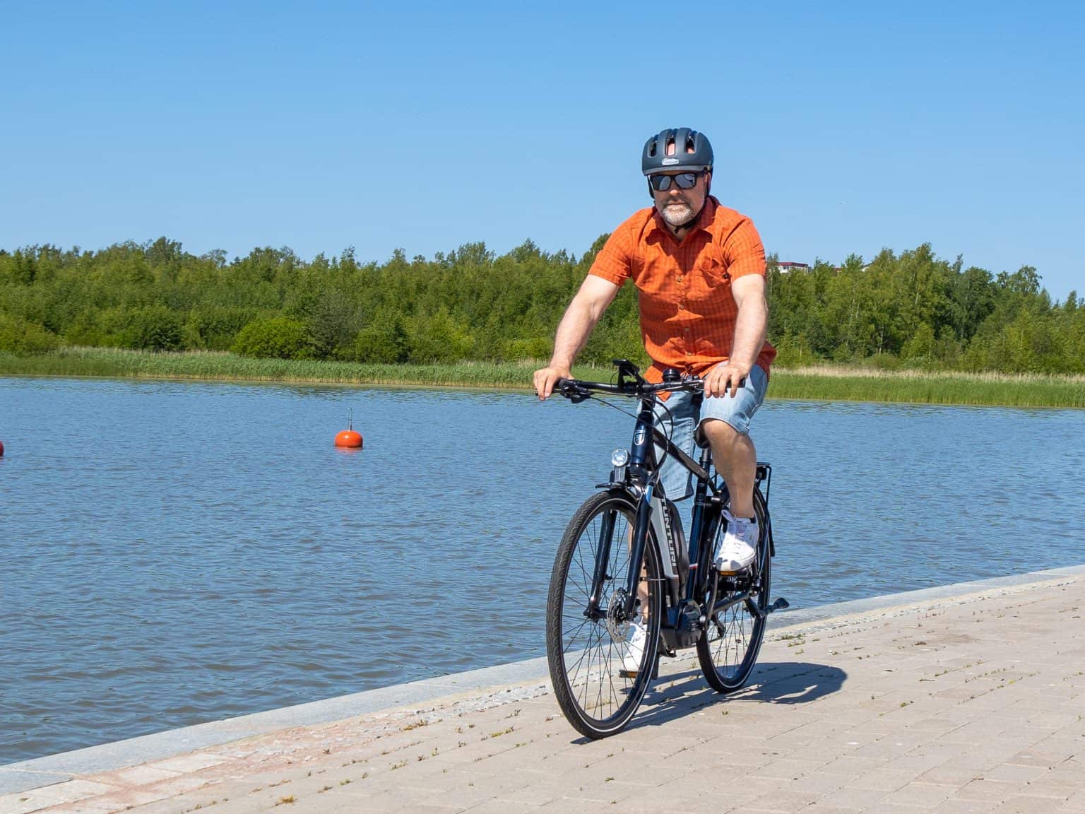 A man wearing a helmet and sunglasses rides a black Tunturi eMotion e-bike along a waterfront path on a sunny day.